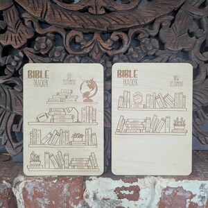 May include: Two wooden Bible tracker boards with a light brown finish. The boards are divided into two sections, one for the Old Testament and one for the New Testament. Each section has a list of books of the Bible with illustrations of books, a globe, a lamp, and a clock.