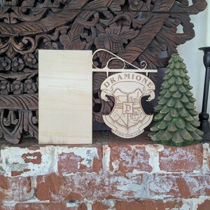 May include: A wooden sign with the words "Dramoine Fanatic Book Club" and a crest featuring a rose and a letter "D". The sign is hanging from a wooden bracket and is displayed on a brick surface.