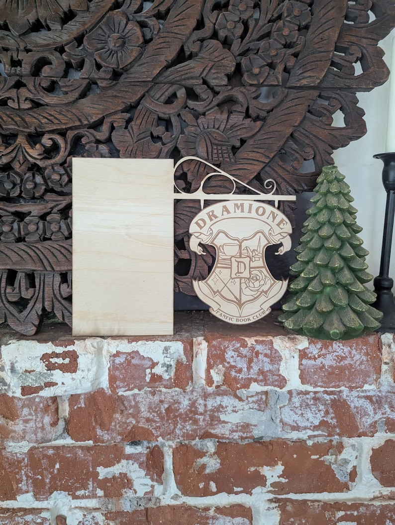 May include: A wooden sign with the words "Dramoine Fanatic Book Club" and a crest featuring a rose and a letter "D". The sign is hanging from a wooden bracket and is displayed on a brick surface.