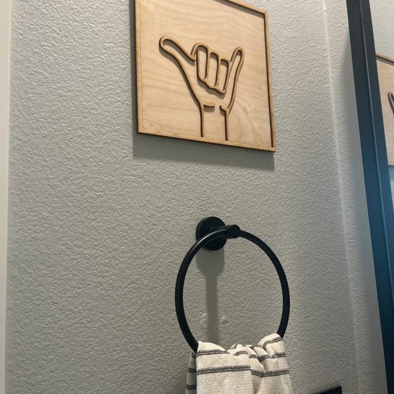 May include: A black metal towel ring with a white and gray striped towel hanging on it. The towel ring is mounted on a light gray wall with a wooden sign with a hand gesture on it.