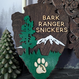 May include: A wooden sign shaped like a park ranger badge with the text "Bark Ranger Snickers" in gold letters. The sign is painted green and brown and features a stylized pine tree, a mountain range, and a paw print.