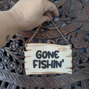May include: A wooden sign with the words "GONE FISHIN'" in black lettering. The sign is light brown with a darker brown border and is suspended by a twine string. The background is a dark brown, carved wooden surface.