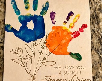 May include: A wooden sign with handprints in blue, orange, green, purple, and yellow paint. The sign says "Grammy" in black lettering and "Thanks for helping us grow!" in brown lettering. Below the handprints is a bouquet of flowers with the names "Teagan" and "Quinn" written on the stems. The sign also says "We love you a bunch!" and "Mother's Day 2023".