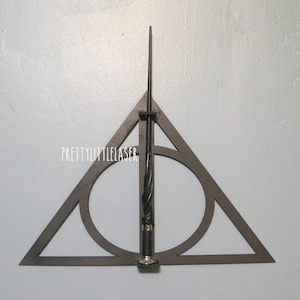 May include: A black metal wall mount in the shape of a triangle with a circle inside, designed to hold a wand. The wand is black with a silver tip and is inserted into the mount.