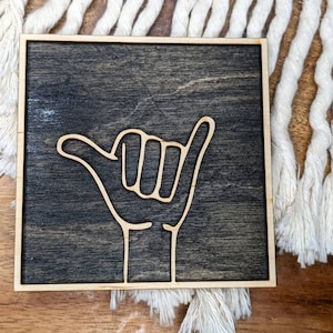 May include: A wooden sign with a dark brown stain and a light wood frame. The sign features a white outline of a hand giving the "hang loose" gesture, also known as the "shaka".