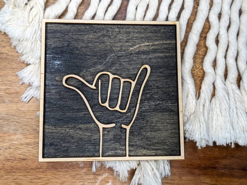 May include: A wooden sign with a dark brown stain and a light wood frame. The sign features a white outline of a hand giving the "hang loose" gesture, also known as the "shaka".