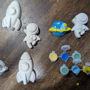 May include: A collection of unpainted ceramic space-themed figurines, including rockets, astronauts, and a planet with a yellow ring. Paint pots with yellow, blue, and green paint are also present, along with a paintbrush. The items are arranged on a wooden surface.
