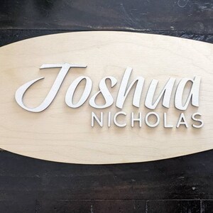 May include: A wooden surfboard-shaped sign with the name "Joshua Nicholas" written in white raised letters.