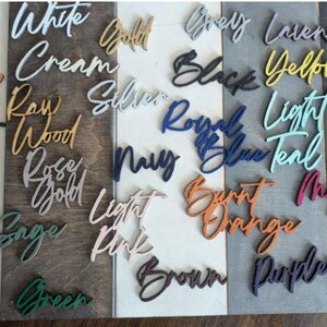 May include: A collection of wooden signs with various color names written on them. The signs are arranged in rows on a wooden surface. The colors include lime green, white cream, gold, grey, lavender, red, yellow, blue, teal, forest green, raw wood, silver, royal blue, light teal, pink, rose gold, navy, burnt orange, magenta, khaki, sage green, light pink, brown, purple, and orange.