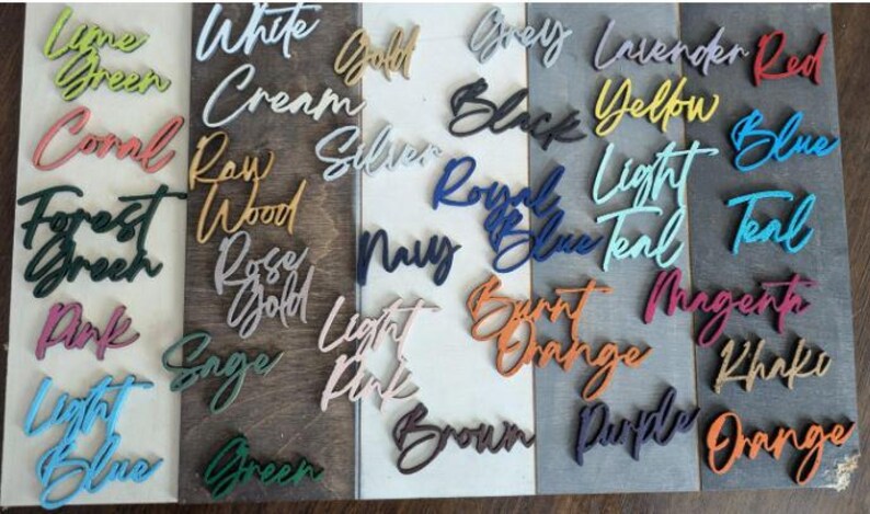 May include: A collection of wooden signs with various color names written on them. The signs are arranged in rows on a wooden surface. The colors include lime green, white cream, gold, grey, lavender, red, yellow, blue, teal, forest green, raw wood, silver, royal blue, light teal, pink, rose gold, navy, burnt orange, magenta, khaki, sage green, light pink, brown, purple, and orange.