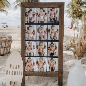 May include: A wooden sign with a brown frame and a white background. The sign has 12 photos of a baby boy in a black and white baseball cap. The photos are arranged in a grid pattern. The sign is on a beach with a surfboard that says "Noah Gabriel" in front of it.