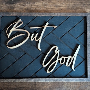 May include: Wooden framed sign with the words "But God" in a cursive font. The background is a dark blue herringbone pattern. The frame is a dark brown color.