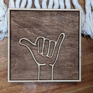 May include: A wooden sign with a white outline of a hand giving the "hang loose" gesture. The sign has a dark brown wood stain.