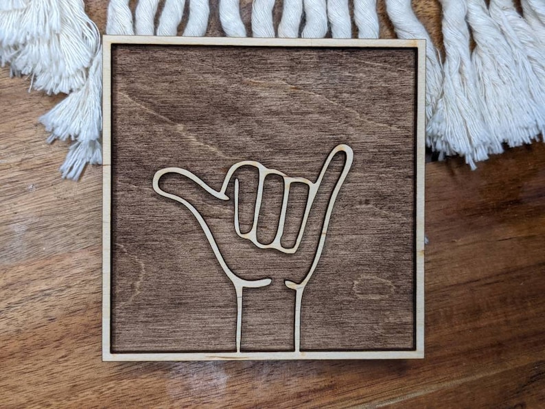 May include: A wooden sign with a white outline of a hand giving the "hang loose" gesture. The sign has a dark brown wood stain.