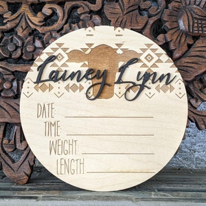 May include: A wooden round plaque with a geometric pattern and a cow silhouette. The plaque has the name "Lainey Lynn" and spaces to record the date, time, weight, and length.