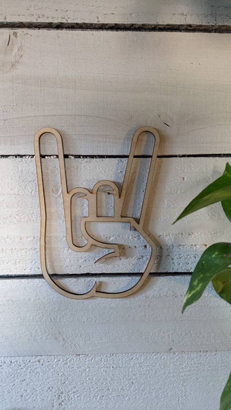 May include: A wooden cutout of a hand making the rock and roll hand gesture. The cutout is light brown and has a smooth finish.