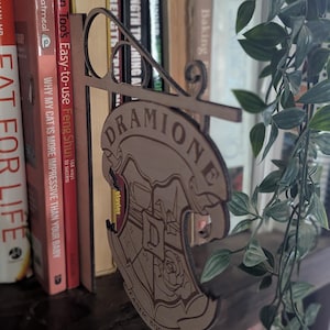 May include: A wooden bookend shaped like a sign with the words "DRAMIONE FANFIC BOOK CLUB" and a Hogwarts house crest. The bookend is brown and has a distressed look.