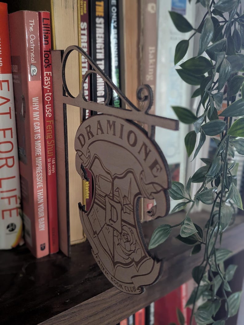 May include: A wooden bookend shaped like a sign with the words "DRAMIONE FANFIC BOOK CLUB" and a Hogwarts house crest. The bookend is brown and has a distressed look.