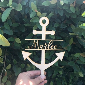 May include: Wooden anchor with the name "Marlee" written on it. The anchor is painted white and is held in a hand against a green leafy background.