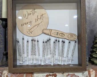 Puede incluir: Un marco de madera con un marco de madera, que contiene un círculo de madera con el texto "Worth every shot" y una ilustración de jeringa. También incluye una prueba de embarazo de madera y varias jeringas.