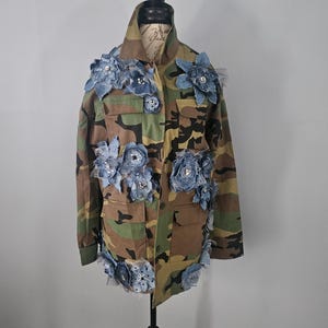 May include: Camouflage jacket adorned with denim flowers and embellishments. The jacket features a classic military-style design with multiple pockets and a button-up front. The denim flowers are in various sizes and add a unique, artistic touch.