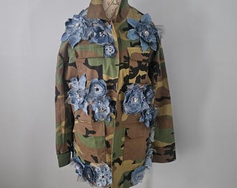 Op de afbeelding: Camouflage jas versierd met denim bloemen en versieringen. De jas heeft een klassiek militair design met meerdere zakken en een knoopsluiting. De denim bloemen zijn in verschillende maten en voegen een unieke, artistieke touch toe.