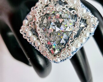 Peut inclure: Une bague carrée avec une base en jean, ornée de chaînes argentées, de perles blanches et de paillettes irisées.