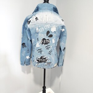 Puede incluir: Una chaqueta vaquera azul claro con detalles desgastados y varios dise&ntilde;os gr&aacute;ficos en blanco y negro, incluyendo calaveras, corazones, ojos y palabras como "Rage", "Bad Girl", "Fierce", "Pain" y "Hurt".