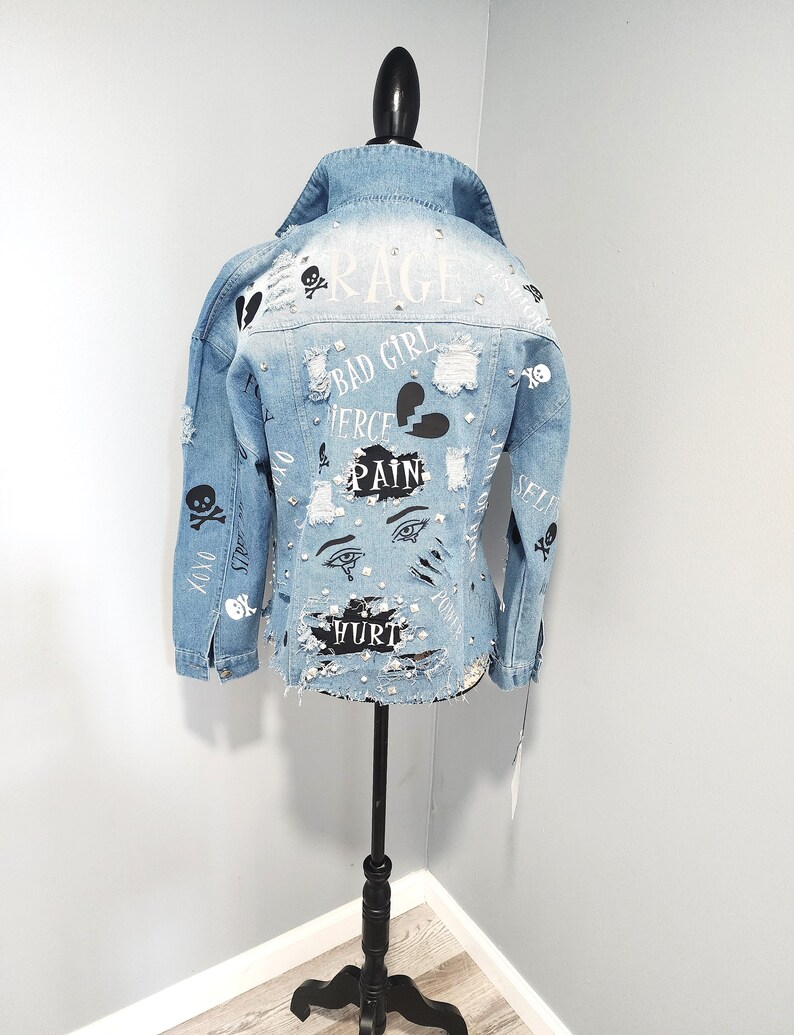 Puede incluir: Una chaqueta vaquera azul claro con detalles desgastados y varios dise&ntilde;os gr&aacute;ficos en blanco y negro, incluyendo calaveras, corazones, ojos y palabras como "Rage", "Bad Girl", "Fierce", "Pain" y "Hurt".