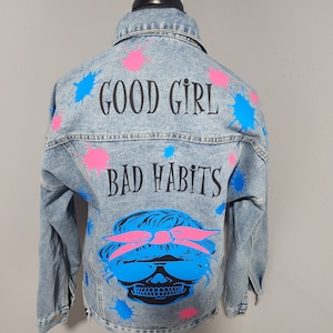 May include: A light blue denim jacket with paint splatters in pink and blue. The jacket has the text "GOOD GIRL BAD HABITS" printed on the back, along with a skull graphic wearing sunglasses and a pink bandana.