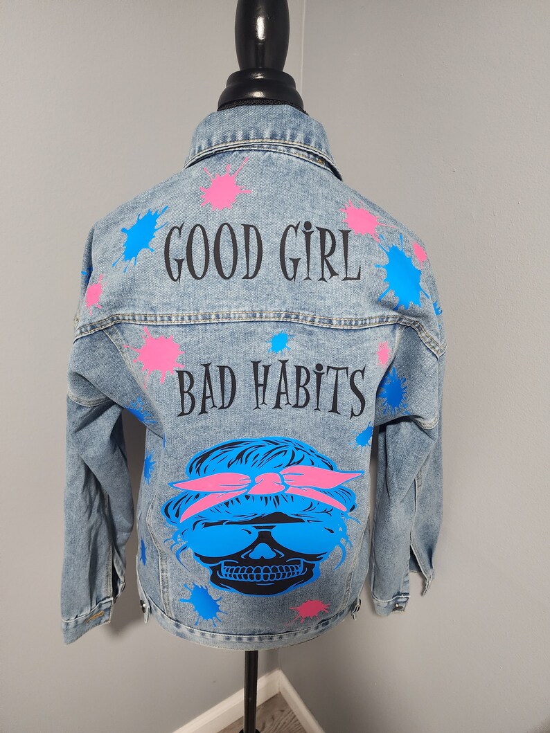 May include: A light blue denim jacket with paint splatters in pink and blue. The jacket has the text "GOOD GIRL BAD HABITS" printed on the back, along with a skull graphic wearing sunglasses and a pink bandana.