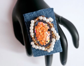 Peut inclure: Une bague avec une grande pierre ovale en résine remplie de paillettes orange. La pierre est entourée d'une bordure de perles blanches et de perles dorées. La bague est posée sur un morceau de tissu en jean bleu.