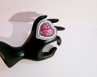 Puede incluir: Un anillo de corazón rosa con un borde de mezclilla y detalles de perlas blancas. El anillo está hecho de lentejuelas y está colocado en un soporte de anillo negro.