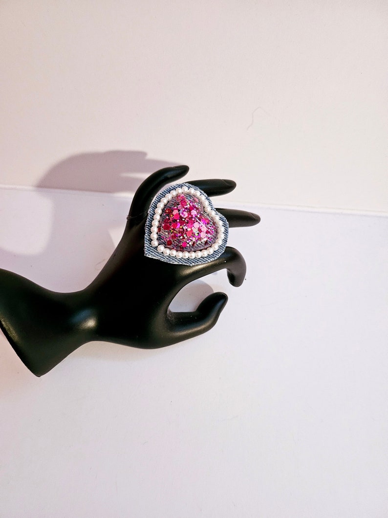 K&ouml;nnte beinhalten: Ein rosafarbener herzf&ouml;rmiger Ring mit einem Jeansrand und wei&szlig;en Perlenakzenten. Der Ring besteht aus Pailletten und befindet sich auf einem schwarzen Ringhalter.
