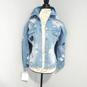Puede incluir: Una chaqueta vaquera azul claro con detalles desgastados, con gr&aacute;ficos de calaveras y huesos cruzados en blanco y negro, pedrer&iacute;a y el texto "FORBIDDEN", "PROMISES" y "STREET"