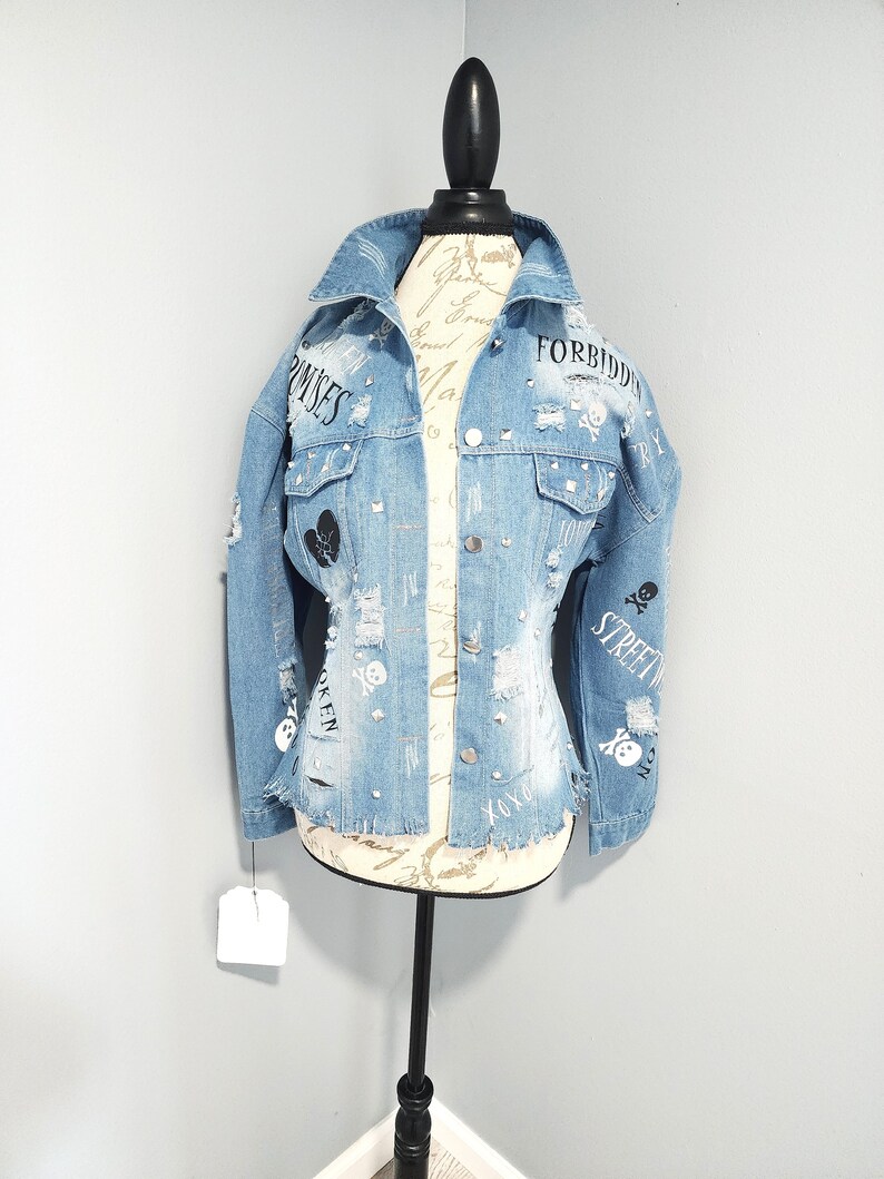 Puede incluir: Una chaqueta vaquera azul claro con detalles desgastados, con gr&aacute;ficos de calaveras y huesos cruzados en blanco y negro, pedrer&iacute;a y el texto "FORBIDDEN", "PROMISES" y "STREET"