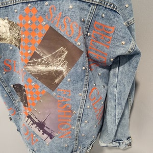 May include: A light blue denim jacket with silver studs and orange text that says "SASSY", "GLAM", "FASHION", and "BELIEVE". The jacket has a patchwork design with images of a bridge, a city skyline, and an airplane.