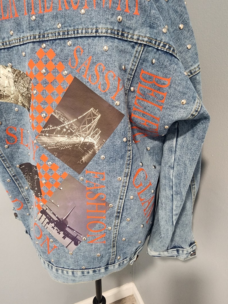 May include: A light blue denim jacket with silver studs and orange text that says "SASSY", "GLAM", "FASHION", and "BELIEVE". The jacket has a patchwork design with images of a bridge, a city skyline, and an airplane.