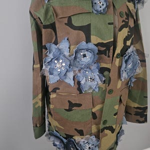 May include: Camouflage jacket adorned with denim and tulle floral embellishments. The jacket features a classic military-style design with multiple pockets and button closures. The blue denim flowers are accented with sparkling rhinestones.