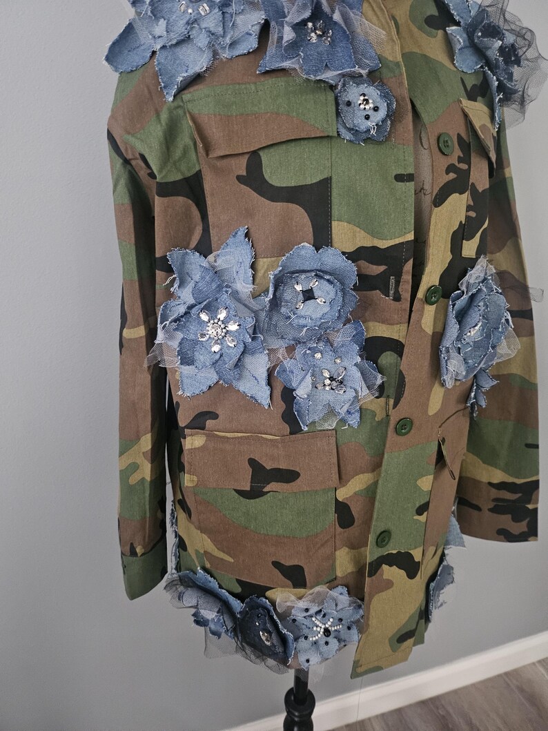 May include: Camouflage jacket adorned with denim and tulle floral embellishments. The jacket features a classic military-style design with multiple pockets and button closures. The blue denim flowers are accented with sparkling rhinestones.