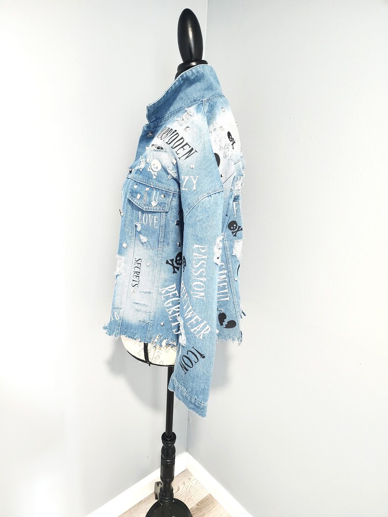 Puede incluir: Chaqueta vaquera azul claro con detalles desgastados, con estampados negros de calaveras y huesos cruzados, corazones y textos, incluyendo "Forbidden", "Love", "Secrets", "Passion", "Gucci", "Recycled Wear" y "Angel".