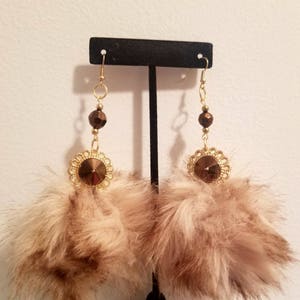 May include: A pair of dangle earrings with brown faux fur pom poms, gold accents, and brown crystal beads.