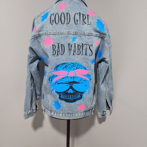 May include: A light blue denim jacket with pink and blue paint splatters. The back of the jacket has the text "GOOD GIRL BAD HABITS" and a skull wearing sunglasses and a pink bandana.