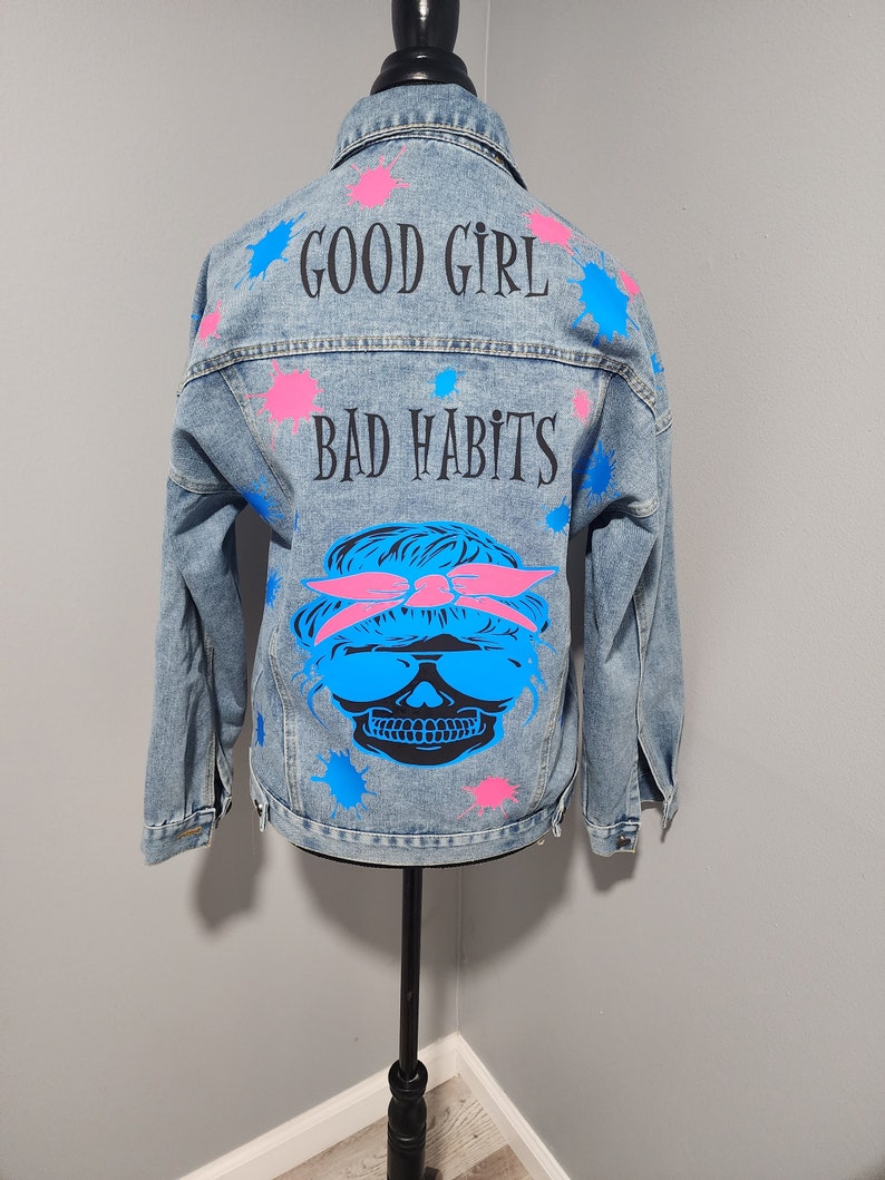 May include: A light blue denim jacket with pink and blue paint splatters. The back of the jacket has the text "GOOD GIRL BAD HABITS" and a skull wearing sunglasses and a pink bandana.