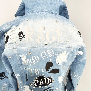 Puede incluir: Chaqueta vaquera azul claro con detalles desgastados, tachuelas y varios estampados gr&aacute;ficos que incluyen "RAGE", "BAD GIRL", "FIERCE", "PAIN" y una calavera con huesos cruzados.