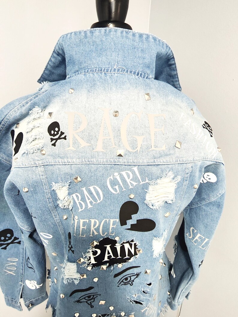 Puede incluir: Chaqueta vaquera azul claro con detalles desgastados, tachuelas y varios estampados gr&aacute;ficos que incluyen "RAGE", "BAD GIRL", "FIERCE", "PAIN" y una calavera con huesos cruzados.