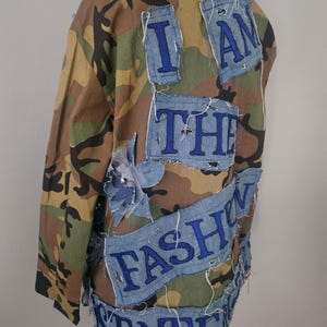 May include: Camouflage jacket with denim patches. The patches are embroidered with the words "I AM THE FASHION STATE" in blue thread. The jacket has a classic military style.