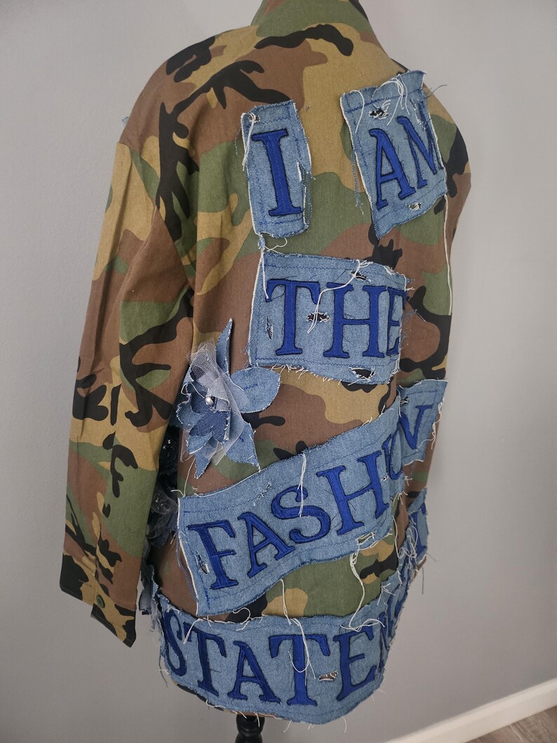 May include: Camouflage jacket with denim patches. The patches are embroidered with the words "I AM THE FASHION STATE" in blue thread. The jacket has a classic military style.