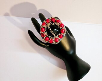 Peut inclure: Une grande bague statement avec une pierre noire entourée d'une chaîne dorée et de perles rouges.