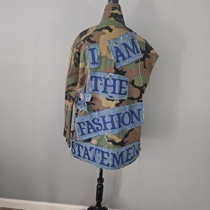 May include: Camouflage jacket with denim patches. The patches spell out "I AM THE FASHION STATEMENT" in blue lettering. The jacket is displayed on a black mannequin stand against a neutral background.